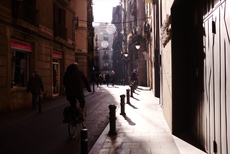 A backlit narrow street in Barcelona's old town, a cyclist and pedestrians silhouetted against warm winter sunlight with festive lights strung overhead.