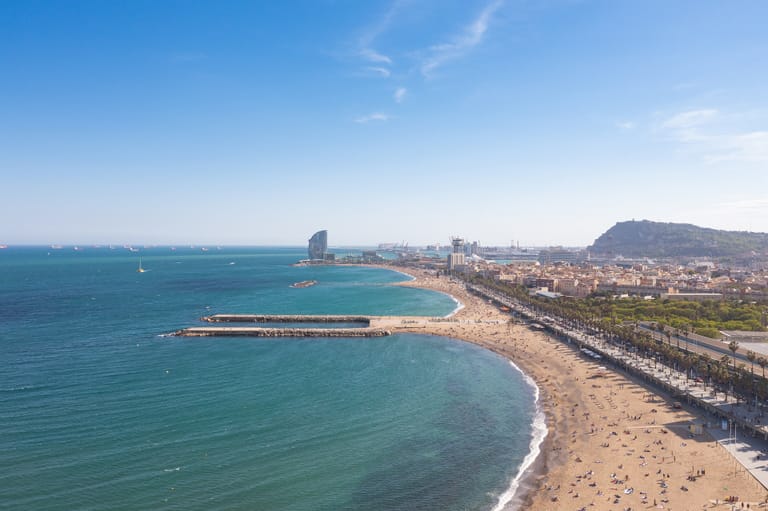 Barcelona's city beach from the air: a long curve of golden sand meeting turquoise water, a palm-lined promenade running beside it and the sail-shaped Hotel Vela at the far end.