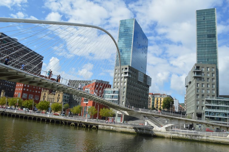 The white Zubizuri footbridge arcing over the Nervión in Bilbao, the Isozaki towers and riverside walk beyond.