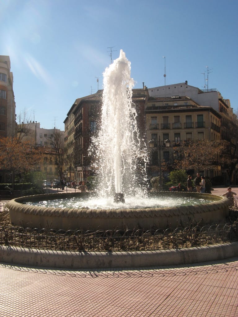 The fountain in Plaza de Olavide, Chamberí, ringed by balconied Madrid apartment blocks in bright winter sun.