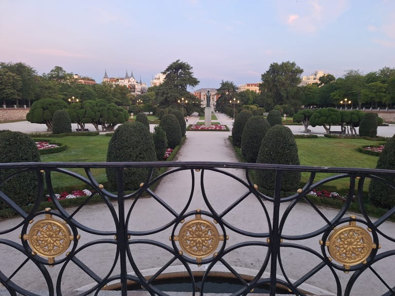 The clipped parterre gardens of El Retiro park in Madrid at dusk, under a pale pink sky.