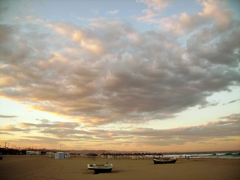 Fishing boats drawn up on the wide sand of Valencia's Malvarrosa beach under a broad evening sky.