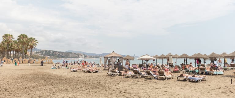 Málaga's La Malagueta beach curving away below the city, parasols and swimmers along the sand with the port beyond.