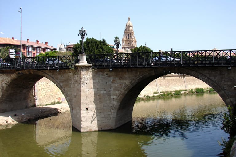 The old stone arches of the Puente de los Peligros over the river Segura in Murcia, the cathedral tower behind.