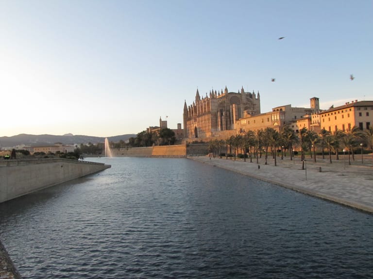 The cathedral of Palma above the still water of the Parc de la Mar, palms and a fountain along the lake in warm evening light.